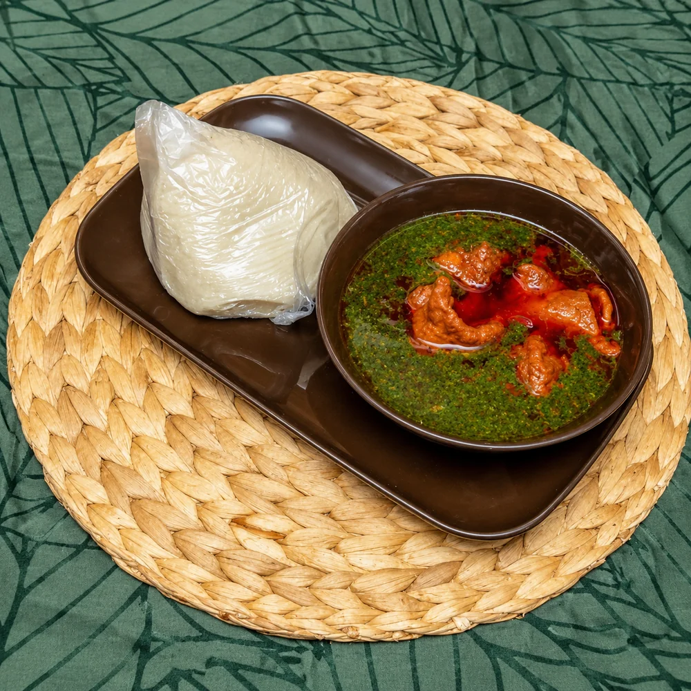 Wrap of white banku on a brown tray beside a bowl of green leaf soup with meat in red palm oil