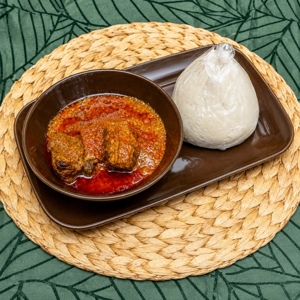 Bowl of okro stew with meat in a rich red sauce, served on a brown tray beside a wrap of banku