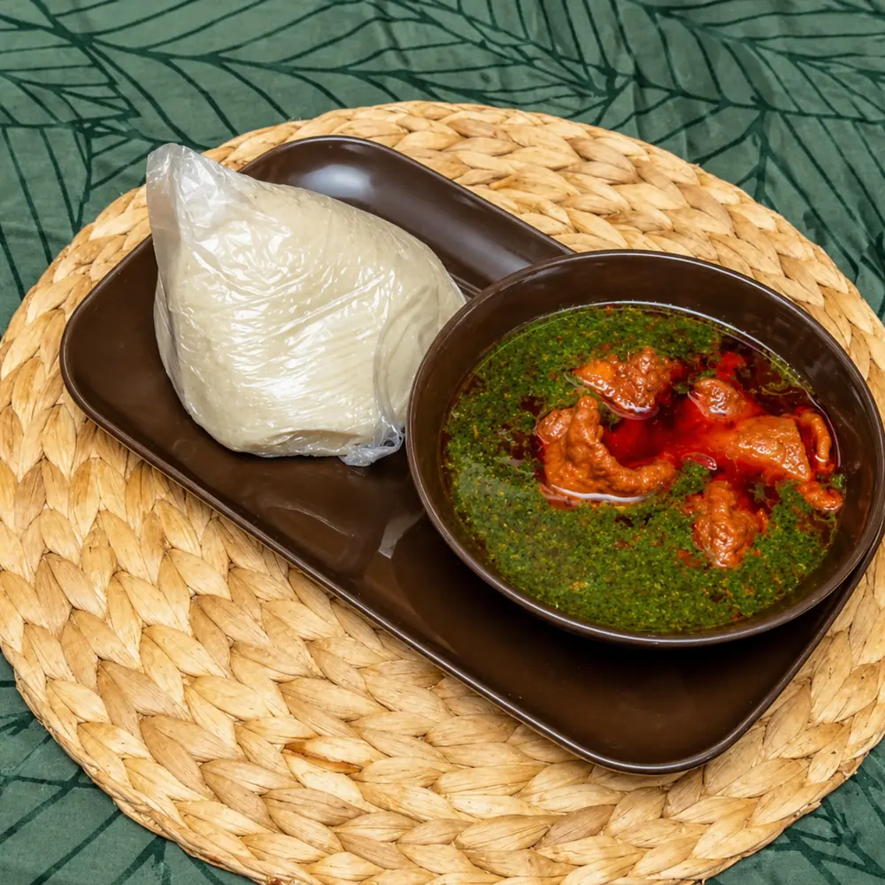 Wrap of white banku on a brown tray beside a bowl of green leaf soup with meat in red palm oil